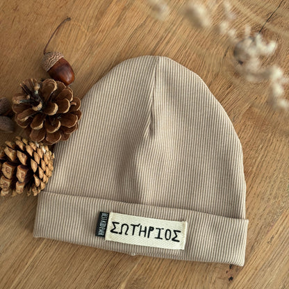 Gerippte Beanie Mütze mit Namen auf weißem Label und shcwarzen Weblabel "Best Kind ever" auf eineem Eicheholztisch daneben Tannenzapfen und Eicheln. Farbe der Mütze: Beige.