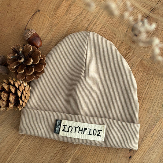 Gerippte Beanie Mütze mit Namen auf weißem Label und shcwarzen Weblabel "Best Kind ever" auf eineem Eicheholztisch daneben Tannenzapfen und Eicheln. Farbe der Mütze: Beige.