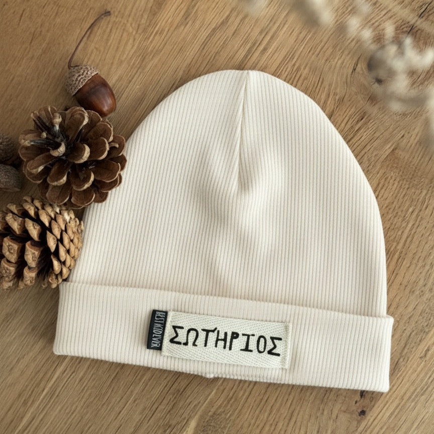 Gerippte Beanie Mütze mit Namen auf weißem Label und shcwarzen Weblabel "Best Kind ever" auf eineem Eicheholztisch daneben Tannenzapfen und Eicheln. Farbe der Mütze: Ecru.