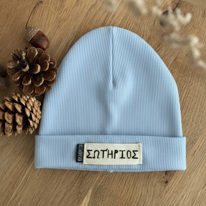 Gerippte Beanie Mütze mit Namen auf weißem Label und shcwarzen Weblabel "Best Kind ever" auf eineem Eicheholztisch daneben Tannenzapfen und Eicheln. Farbe der Mütze: Hellblau.