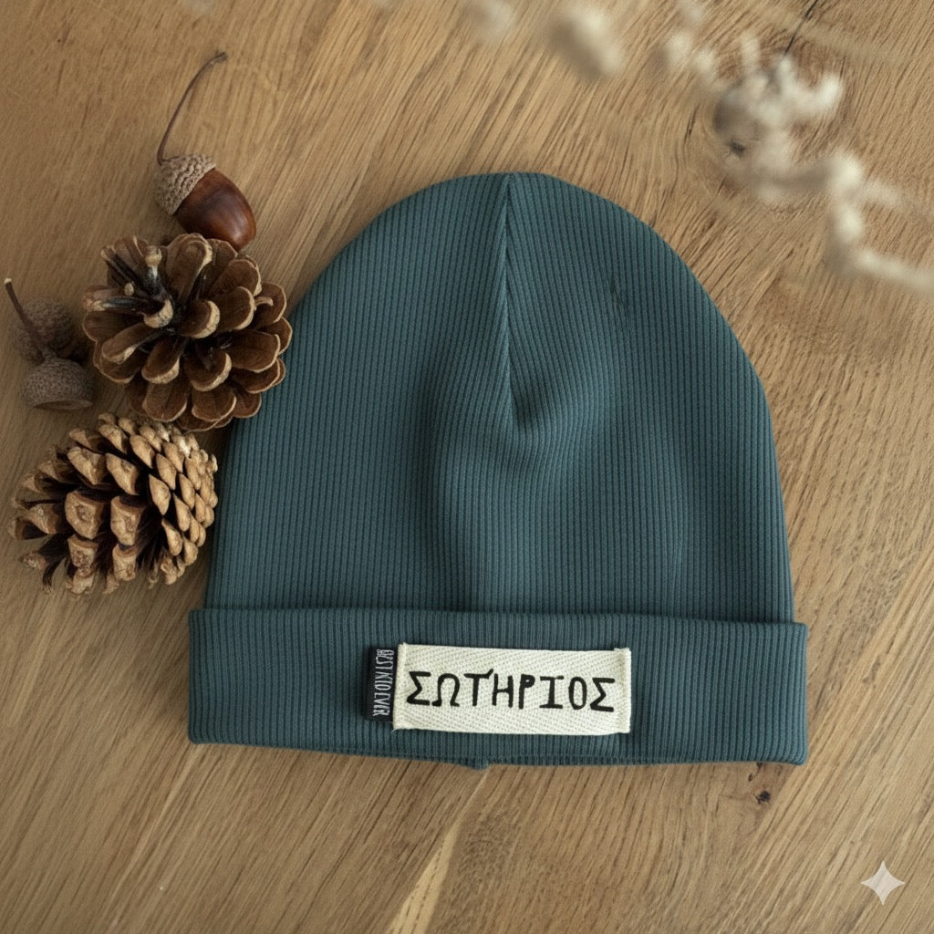 Gerippte Beanie Mütze mit Namen auf weißem Label und shcwarzen Weblabel "Best Kind ever" auf eineem Eicheholztisch daneben Tannenzapfen und Eicheln. Farbe der Mütze: Petrol.